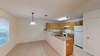 Located at 7791 Lilah Ln in Pensacola, Florida, this two-story home offers a practical and comfortable layout designed for everyday living. The kitchen serves as a focal point of the main level, featuring warm honey-toned wood cabinetry that provides generous storage, granite-style countertops, and a peninsula with a built-in sink that opens toward the adjoining living area. White appliances, including a refrigerator and electric range, complement the clean, bright aesthetic of the space. A decorative pendant light hangs above the peninsula, adding a subtle design touch to the open layout. Light-toned ceramic tile flooring runs throughout the main level, creating a cohesive and easy-to-maintain surface underfoot. The open-concept design between the kitchen and living area encourages a natural flow, making the space feel connected and functional. Recessed and flush-mounted ceiling fixtures provide ample overhead lighting throughout. Neutral white walls keep the interior feeling bright and versatile, ready to accommodate a variety of décor styles. Spanning two floors, this Pensacola home offers room to spread out, with the upper level providing additional living or sleeping space to suit the needs of its residents.