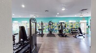 This fitness center is a thoughtfully designed, single-floor facility that balances energetic aesthetics with a comprehensive range of exercise equipment. The space is immediately distinguished by its bold visual identity — a striking teal accent wall adorned with a large, multicolored geometric mural rendered in blues, greens, yellows, and grays creates a dynamic and motivating atmosphere throughout.

The flooring consists of durable wood-look planks that span the entire space, offering both a clean appearance and easy maintenance. Recessed lighting and ceiling fans provide consistent illumination and air circulation, contributing to a comfortable workout environment.

The cardio zone is anchored along one windowed wall, where multiple large windows allow natural light to fill the area and offer outdoor views. This section houses treadmills, an elliptical trainer, and a recumbent stationary bike, with wall-mounted flat-screen televisions positioned above for entertainment during workouts.

The strength training area is generously equipped with multiple cable and multi-functional resistance machines positioned on protective floor mats. A full dumbbell rack lines the wall alongside the machines, catering to a variety of free-weight exercises. Large mirrors are mounted along one wall, enhancing the sense of space and allowing for proper form monitoring.

Fitness center rules are posted near the entrance, and the overall layout ensures clear pathways and ample room between equipment stations.