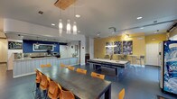 This energetic single-floor community clubhouse, associated with Campus Evolution Villages, is designed to foster social interaction and recreation. The space is anchored by an open-plan layout with polished concrete flooring throughout, lending a modern, cohesive feel.

The entry lounge greets visitors with a distinctive curved, corrugated-panel seating pod, an artistic cluster of Edison bulb pendant lights overhead, and a large chalkboard wall displaying "Live Big." A sliding barn door adds an industrial-chic touch nearby.

Adjacent to the entry is a cozy sitting area bathed in warm orange tones, featuring a curved pink sofa, striped upholstered armchairs, and a large framed artwork. A full-panel glass garage-style door floods the room with natural light and offers views of the exterior stone facade.

The central dining and recreation zone features long communal tables with vibrant orange chairs, alongside a full-size pool table set against a bold yellow accent wall adorned with large-scale black-and-white sports photography. Track lighting highlights the athletic artwork above.

The kitchen area is well-appointed with wrap-around countertops, rich wood cabinetry, a barn door, pendant lighting, and a bold navy blue accent wall featuring a welcoming chalkboard sign. Appliances including a microwave and beverage cooler make the space fully functional for community gatherings.