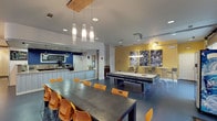 This energetic single-floor community clubhouse, associated with Campus Evolution Villages, is designed to foster social interaction and recreation. The space is anchored by an open-plan layout with polished concrete flooring throughout, lending a modern, cohesive feel.

The entry lounge greets visitors with a distinctive curved, corrugated-panel seating pod, an artistic cluster of Edison bulb pendant lights overhead, and a large chalkboard wall displaying "Live Big." A sliding barn door adds an industrial-chic touch nearby.

Adjacent to the entry is a cozy sitting area bathed in warm orange tones, featuring a curved pink sofa, striped upholstered armchairs, and a large framed artwork. A full-panel glass garage-style door floods the room with natural light and offers views of the exterior stone facade.

The central dining and recreation zone features long communal tables with vibrant orange chairs, alongside a full-size pool table set against a bold yellow accent wall adorned with large-scale black-and-white sports photography. Track lighting highlights the athletic artwork above.

The kitchen area is well-appointed with wrap-around countertops, rich wood cabinetry, a barn door, pendant lighting, and a bold navy blue accent wall featuring a welcoming chalkboard sign. Appliances including a microwave and beverage cooler make the space fully functional for community gatherings.