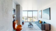 This contemporary high-rise residence showcases a refined modern aesthetic defined by clean lines, an open layout, and an abundance of natural light. The living area serves as the centerpiece of the space, anchored by dramatic floor-to-ceiling windows that frame sweeping panoramic city views. Exposed concrete columns add an industrial character that contrasts elegantly with the crisp white walls and light hardwood flooring found throughout. The living room comfortably accommodates a full seating arrangement and media setup, with a sleek TV console positioned against one of the concrete columns.

Adjacent to the living area, a dedicated dining space features high ceilings and offers a bright, airy setting ideal for everyday meals or entertaining. The kitchen is outfitted with light blue cabinetry, stainless steel appliances including a gas range and over-the-range microwave, and a chevron-patterned tile backsplash with under-cabinet lighting.

The bedroom is generously proportioned with its own floor-to-ceiling window wall offering elevated city views, a built-in closet, and light hardwood flooring. The bathroom features large-format tile flooring, a floating vanity with an integrated sink, and a full mirror above.

Throughout the residence, the interplay of concrete, glass, and light wood creates a cohesive, sophisticated atmosphere with a distinctly urban character.