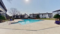 This community amenity space is centered around a generously sized outdoor swimming pool, finished with classic brick-edged coping and filled with clear, blue water. The pool area is surrounded by a broad concrete sundeck that provides ample space for lounging and outdoor activities. Entry points into the pool are accessible via stainless steel handrails, and the deck is enclosed by a wrought iron perimeter fence, ensuring both safety and a defined boundary for the area.

Adjacent to the pool sits a single-story clubhouse structure featuring a low-pitched gabled roofline with dark trim accents and multiple chimney details that complement the architectural style of the surrounding residential buildings. The clubhouse includes a glassed sunroom-style section that allows natural light to fill the interior, as well as a restroom facility finished with white shiplap-style wall panels, dark door and trim accents, and transom windows that bring in additional daylight. Mature trees and neatly trimmed shrubbery frame the space, adding natural character. The multi-building residential complex visible in the background features two-story structures with balconies and matching dark-trimmed architecture, creating a cohesive visual environment throughout the community.