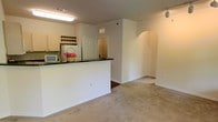 This single-floor residence offers a well-designed, open-concept layout that seamlessly connects the kitchen and living areas. The kitchen is equipped with light wood cabinetry, a white refrigerator, and a raised bar counter finished with dark granite-style countertops, providing both functionality and a natural dividing point between the cooking and living spaces. A built-in wine rack adds a thoughtful storage detail above the cabinetry. The living area features warm, neutral wall tones and plush carpeting underfoot, creating a comfortable and inviting atmosphere throughout. Crown molding runs along the ceiling, adding a touch of classic architectural refinement. A standout design element is the elegant arched doorway on the far wall, which lends character and visual interest to the space. Track lighting is installed along the ceiling, offering adjustable illumination to complement the recessed fluorescent fixtures in the kitchen. Clean white baseboards frame each room, and the overall color palette — soft beiges and creams — contributes to a bright and cohesive feel throughout the home.