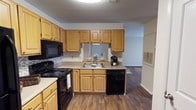 This single-floor residence offers a comfortable and practical layout designed to maximize everyday functionality. The kitchen serves as a focal point of the home, showcasing warm honey-toned wood cabinetry that provides generous storage space both above and below the countertops. Black appliances — including a range, built-in microwave, and dishwasher — add a sleek contrast against the light countertops and cabinetry. A double-basin sink is positioned beneath a window, allowing natural light to filter into the space and brighten the work area. A backsplash accent adds a subtle decorative touch to the kitchen's clean, practical aesthetic. Rich hardwood-style flooring runs throughout, lending warmth and visual continuity from the kitchen into the adjoining living areas. A textured ceiling adds dimension overhead. The open sightline from the kitchen into the neighboring room creates a connected, airy feel throughout the home. Overall, the space presents a well-maintained interior with a neutral palette, offering a welcoming and move-in-ready environment suited for comfortable day-to-day living.