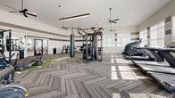 Located at The One at Hope Mills in Fayetteville, NC, this single-floor fitness center encompasses a generous and thoughtfully designed exercise facility divided into two distinct exercise rooms. The main exercise area immediately impresses with soaring ceilings, large windows that flood the space with natural light, and a sleek herringbone-patterned carpet. Overhead, modern linear pendant lights and ceiling fans maintain a comfortable, well-lit environment throughout. A branded turf runner anchored with The One at Hope Mills logo runs through the center of the main floor, adding a bold, community-centric design element. This primary room features a comprehensive array of equipment, including a multi-functional cable machine, a heavy punching bag, a mini trampoline, weight benches, dumbbells, barbells, and stair climbers. The walls are accented with distinctive horizontal stripes in neutral tones of white, taupe, and green. A second exercise room, finished with rich hardwood flooring, is dedicated to functional training, housing a branded multi-rack system with TRX straps, medicine balls, and an additional punching bag, along with a ceiling fan and full-length mirrors to support form and technique. Together, the two rooms deliver a comprehensive, gym-quality fitness experience.