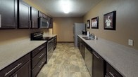This single-floor community room offers a well-appointed gathering space designed for comfort and functionality. The fully equipped galley-style kitchen is outfitted with dark espresso cabinetry, light stone-toned countertops, stainless steel appliances including a range, microwave, refrigerator, and dishwasher, all set atop durable tile flooring. The kitchen opens directly into a dedicated dining area, furnished with a dark wood table and seating for six, accented by warm neutral walls and carpeted flooring.

The expansive lounge area serves as the centerpiece of the community room, featuring soaring vaulted ceilings with exposed beam detailing, track lighting, and a classic fireplace with a dark mantle. A wall-mounted television completes the entertainment setup. Large sliding glass doors and wide windows flood the space with natural light while providing direct access to an outdoor deck, offering views of the surrounding landscape. Additional seating areas throughout the lounge create versatile spaces for both small gatherings and larger group events. The overall ambiance is warm and inviting, combining practical amenities with an open, airy atmosphere suited for community gatherings.