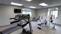 This single-level fitness room is designed to support a well-rounded exercise routine in a clean, functional environment. The space features a drop ceiling with recessed panel lighting that provides bright, even illumination throughout, complemented by a window that allows natural light to filter in, creating an open and inviting atmosphere.

The room is outfitted with a selection of cardio equipment, including treadmills positioned toward the front of the space, making them easily accessible. A wall-mounted flat-screen television is positioned at a comfortable viewing height, ideal for entertainment during workouts. Toward the rear of the room, a variety of white strength and resistance machines are arranged neatly, offering users options for targeted muscle training and rehabilitation-style exercises.

The flooring consists of low-pile patterned carpet, which adds a degree of comfort and sound absorption. The neutral gray walls contribute to a calm, focused atmosphere. A seating area with chairs is available along the perimeter, providing a convenient rest area. Overall, the space is thoughtfully arranged to maximize usability while maintaining an uncluttered, professional appearance.