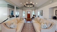 This lobby makes a strong first impression with its soaring vaulted ceilings that lend an immediate sense of openness and grandeur. The architectural design combines classic and coastal influences, highlighted by white shiplap wainscoting that lines the walls and is complemented by soft sage-green paint above. Rich, dark hardwood floors run throughout, providing a warm contrast to the light, neutral color palette.

A striking wrought-iron candle-style chandelier serves as a central focal point, suspended beneath the peak of the vaulted ceiling and anchoring the seating area below. Two generously sized cream sofas face each other across a bold geometric-patterned area rug, creating an inviting and symmetrical lounge arrangement. Accent chairs and console tables with decorative lamps are thoughtfully placed throughout, adding both function and style.

Natural light flows in abundantly through multiple windows dressed in flowing white drapery, as well as through a wood-framed glass entry door flanked by side windows. A built-in coffee station is tucked into a recessed nook along one wall, adding a practical amenity. Curated artwork and decorative mirrors complete the refined, welcoming atmosphere of this well-appointed lobby.