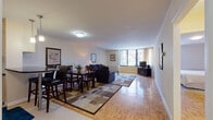 This single-floor residence offers a thoughtfully arranged layout with a seamless flow between its main living areas. The open-plan living and dining space is generously sized, anchored by classic parquet hardwood flooring that runs throughout much of the unit. Large windows in the living area allow ample natural light to fill the space while providing outdoor views, creating a bright and welcoming ambiance. A dedicated dining area accommodates a full table and chair set, positioned conveniently adjacent to the kitchen.

The kitchen is well-equipped with stainless steel appliances, including a refrigerator, gas range, microwave, and dishwasher. White cabinetry provides generous storage, complemented by dark countertops and a tiled floor. A breakfast bar with seating adds a casual dining option, connecting the kitchen to the main living area.

The bedroom is comfortably proportioned, featuring the same parquet flooring, soft neutral walls, and a window that brings in natural light. It offers ample space for furnishings and includes closet access. The bathroom features large-format tile flooring, a warm wood vanity with a granite countertop, a mirrored medicine cabinet, and bright overhead lighting, presenting a clean and functional design throughout.