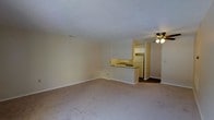 This single-floor residential space features a practical and functional layout designed to maximize everyday living comfort. The main living area is generously sized, finished with wall-to-wall carpet flooring in a neutral tone that complements the soft, cream-colored walls throughout. A textured popcorn ceiling adds a classic touch to the space, while a ceiling fan with a built-in light fixture provides both air circulation and ambient lighting.

The kitchen is positioned in an open-concept arrangement, separated from the living area by a peninsula countertop that doubles as a breakfast bar or prep space. White cabinetry lines the upper walls of the kitchen, offering ample storage, while a white refrigerator and a sink with a dark countertop complete the functional setup. The kitchen floor transitions from the carpeted living area to a darker hard flooring surface, clearly delineating the two zones.

The overall ambiance of the space is clean, understated, and move-in ready, with a neutral palette that makes it easy to personalize with furnishings and décor. The open flow between the living and kitchen areas creates a cohesive, connected feel throughout the single-floor layout.