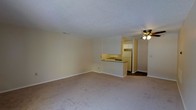 This single-floor residential space features a practical and functional layout designed to maximize everyday living comfort. The main living area is generously sized, finished with wall-to-wall carpet flooring in a neutral tone that complements the soft, cream-colored walls throughout. A textured popcorn ceiling adds a classic touch to the space, while a ceiling fan with a built-in light fixture provides both air circulation and ambient lighting.

The kitchen is positioned in an open-concept arrangement, separated from the living area by a peninsula countertop that doubles as a breakfast bar or prep space. White cabinetry lines the upper walls of the kitchen, offering ample storage, while a white refrigerator and a sink with a dark countertop complete the functional setup. The kitchen floor transitions from the carpeted living area to a darker hard flooring surface, clearly delineating the two zones.

The overall ambiance of the space is clean, understated, and move-in ready, with a neutral palette that makes it easy to personalize with furnishings and décor. The open flow between the living and kitchen areas creates a cohesive, connected feel throughout the single-floor layout.