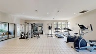 Located at 117 SE 16th Ave in Gainesville, Florida, this fitness center offers a comprehensive exercise facility across a single, open floor plan. The space is finished with clean white tile flooring throughout the main exercise area, transitioning to carpet in the dedicated strength and free-weight zone, providing practical surface variety for different workout activities.

The facility is well-lit by recessed ceiling lights and benefits from abundant natural light streaming through large multi-pane windows that frame views of the surrounding outdoor greenery. Two sets of glass-paneled double doors provide convenient access points, connecting the interior to the outdoor areas and enhancing the sense of openness.

The cardio section is generously stocked with treadmills, stationary bikes, and elliptical trainers, each equipped with individual monitors. The strength training area features a multi-station cable machine with bench, a power tower for bodyweight exercises, and a rack of medicine balls for functional training. A wall-mounted flat-screen television is positioned for viewing during workouts. Additional amenities include a water dispenser with a supply of water jugs, ensuring users stay hydrated. Mirrors on one wall expand the sense of space and support form monitoring. The overall ambiance is clean, functional, and well-maintained, making it a practical and inviting environment for a full range of fitness activities.
