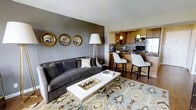 This single-floor space showcases an open-concept design that seamlessly connects the living area and kitchen, creating a cohesive and functional layout. The living room is anchored by a plush charcoal gray sofa positioned against a bold dark gray accent wall, which serves as a striking focal point. Three decorative gold-framed convex mirrors are arranged above the sofa, adding an elegant and curated aesthetic. A patterned area rug in neutral tones with soft blue and brown accents grounds the seating area, while a sleek white coffee table with a chrome base completes the arrangement.

A tall brass floor lamp provides warm ambient lighting, complementing the overall color palette. The kitchen is directly accessible from the living space and features warm wood cabinetry, dark granite countertops, and a mosaic tile backsplash that adds texture and visual interest. Stainless steel appliances, including a refrigerator and range, contribute a modern edge. A raised breakfast bar with white upholstered counter-height chairs offers casual seating. Pendant lighting above the bar and under-cabinet illumination enhance the kitchen's functionality. Hardwood flooring flows throughout, tying the spaces together cohesively.