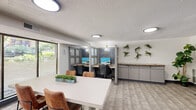 Located at 8750 First Ave in Silver Spring, MD, this business center suite offers a well-organized, single-floor workspace designed to accommodate a variety of professional needs. The layout balances collaborative and focused work areas within a compact yet efficiently utilized footprint.

A central conference-style table with leather chairs serves as the primary meeting space, positioned directly in front of expansive floor-to-ceiling windows that flood the area with natural light and offer views of the surrounding tree-lined exterior. Frosted glass panel partitions and a sliding glass door delineate the suite from adjacent spaces while maintaining an open, airy feel.

The workstation area features individual cubicle-style desks separated by frosted glass and gray-paneled dividers, offering a degree of privacy without fully enclosing the space. Built-in cabinetry along one wall provides practical storage, while decorative wall-mounted plants and a framed art piece contribute warmth and character to the otherwise modern, neutral interior.

Flooring throughout combines patterned carpet tile in the meeting zone with wood-look vinyl in the workstation area. Circular flush-mount ceiling lights provide consistent overhead illumination. The textured ceiling, gray baseboard trim, and clean white walls complete a professional and functional aesthetic suited for a business or office environment.