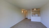 This single-floor unit presents an open-concept layout that connects the main living area to the kitchen. The living space is finished with wall-to-wall carpet in a neutral beige tone, contributing to a simple and understated aesthetic throughout. Walls are painted in a light, off-white hue, and the textured ceiling adds a subtle architectural detail.

The kitchen is partially open to the living area, separated by a half-wall partition that creates a pass-through effect. This design allows visual connectivity between the two spaces while maintaining a degree of separation. The kitchen features light wood-toned cabinetry, a countertop workspace, and standard appliances, including what appears to be a refrigerator and range. Overhead cabinetry provides practical storage space.

Lighting in the unit is provided by wall-mounted sconces and a ceiling-mounted fixture near the kitchen, offering ambient illumination throughout. A hallway visible toward the back of the unit likely leads to additional rooms. The overall ambiance of the space is clean and functional, with a neutral palette that offers a versatile foundation for various interior arrangements.