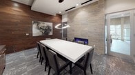 This professionally designed conference room offers a refined and functional environment suited for meetings and collaborative gatherings. At the center of the space sits a substantial white conference table surrounded by sleek, dark leather chairs, providing seating for multiple attendees. Overhead, a contemporary circular pendant light fixture serves as a focal point, complemented by recessed ceiling lighting that ensures even illumination throughout.

One wall features a striking combination of large-format stone-look tile cladding and a linear built-in fireplace with blue accent lighting, adding warmth and a distinctive design element to the room. Adjacent to this, a rich wood-paneled accent wall displays a bold piece of abstract artwork, lending the space a gallery-like quality. A wall-mounted flat-screen display and a dark wood credenza with ample storage provide practical amenities along another wall.

The room is enclosed by frosted glass sliding partition doors, offering privacy while maintaining a visual connection to adjacent spaces. Patterned carpet tile in neutral tones grounds the interior with texture and depth. The overall design balances professional functionality with upscale finishes, creating an impressive setting for business engagements.
