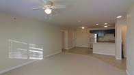 This single-floor space features an open-concept layout that seamlessly connects the living area to the kitchen, creating a spacious and functional environment. The living area is finished with plush neutral-toned carpeting and painted in a soft beige palette, lending a warm and inviting ambiance throughout. A ceiling fan with an integrated light fixture serves as a central overhead feature, providing both airflow and ambient lighting to the main living space. Recessed lighting is also present, offering additional illumination across the open floor plan.

The kitchen is positioned toward the rear of the space and is outfitted with dark cabinetry that provides a strong contrast against the lighter surroundings. A stainless steel refrigerator and a peninsula-style counter are notable features, with the counter offering convenient workspace and potential for seating. The kitchen area transitions from carpet to a hard surface flooring, clearly delineating the two functional zones.

Two interior doorways are visible along the back wall, suggesting access to additional rooms such as a bedroom or storage areas. The space receives natural light through windows, as evidenced by bright reflections on the floor.