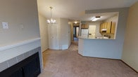 This single-floor space showcases an open-concept design that seamlessly connects the living area and kitchen. The living room is anchored by a tile-surround fireplace with a white mantel, adding a functional focal point to the space. Overhead, a classic chandelier with candle-style lights enhances the room's character, while neutral-toned carpet extends throughout the main living area, creating a warm, cohesive feel.

The kitchen features light wood cabinetry, a curved peninsula with a raised bar counter, and essential appliances including a white refrigerator and electric range. A ventilation hood is mounted above the cooktop, and under-cabinet lighting adds practical illumination to the workspace. Tile flooring in the kitchen area provides a clean transition from the carpeted living space.

A bifold-door closet offers convenient storage near the living area, while a hallway leads toward additional rooms and an entry door beyond. The vaulted ceiling line and open sightlines contribute to an airy, spacious ambiance throughout the main living areas. Overall, the layout is practical and well-connected, suited for comfortable everyday living.