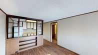 This single-floor residence offers a practical and functional layout that connects the living and kitchen areas in an open, airy fashion. The kitchen is partially separated from the living space by a half-wall partition featuring built-in shelving with warm wood-toned finishes, creating a sense of openness while maintaining a defined cooking area. The kitchen itself is equipped with white cabinetry, open shelving, a white range, and a refrigerator, with a light-colored countertop and a sink positioned along the partition wall. The living area features durable hardwood-style flooring in a rich, warm brown tone that extends throughout the space, complementing the wood accents of the cabinetry and trim. Neutral beige walls and white textured ceilings with a classic popcorn finish contribute to a clean and understated aesthetic. Dark brown crown molding and baseboards provide a defined contrast against the lighter walls. A hallway visible from the main living area leads to additional rooms, including a bathroom, offering a well-connected and efficiently designed floor plan. Overall, the space presents a cohesive, warm interior with functional design elements suited to everyday living.