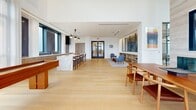 Located at 4909 Auburn Ave in Bethesda, MD, this refined single-floor space blends modern design with upscale functionality across two distinct living areas. Light hardwood flooring runs seamlessly throughout, unifying the open layout and complementing the bright, airy atmosphere created by an abundance of natural light.

The main gathering area features soaring double-height ceilings, expansive floor-to-ceiling windows, and direct access to an outdoor terrace with sweeping views of the surrounding Bethesda skyline. A dramatic marble-clad fireplace with integrated media display serves as a focal point, flanked by elegant seating arrangements. An intricate geometric metal screen partition adds architectural interest while defining zones within the open plan. Built-in shelving and a decorative pendant light enhance the curated aesthetic.

A second living area showcases exposed wood beam ceiling details, a second marble fireplace, and generous window walls that flood the space with natural light. Adjacent to this is an open kitchen and lounge zone featuring a substantial marble waterfall island with bar seating, warm wood cabinetry, and industrial-style pendant lighting. A dedicated game area with a hardwood shuffleboard table rounds out the space, offering a versatile and inviting environment suited for both relaxation and entertainment.
