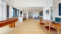 Located at 4909 Auburn Ave in Bethesda, MD, this refined single-floor space blends modern design with upscale functionality across two distinct living areas. Light hardwood flooring runs seamlessly throughout, unifying the open layout and complementing the bright, airy atmosphere created by an abundance of natural light.

The main gathering area features soaring double-height ceilings, expansive floor-to-ceiling windows, and direct access to an outdoor terrace with sweeping views of the surrounding Bethesda skyline. A dramatic marble-clad fireplace with integrated media display serves as a focal point, flanked by elegant seating arrangements. An intricate geometric metal screen partition adds architectural interest while defining zones within the open plan. Built-in shelving and a decorative pendant light enhance the curated aesthetic.

A second living area showcases exposed wood beam ceiling details, a second marble fireplace, and generous window walls that flood the space with natural light. Adjacent to this is an open kitchen and lounge zone featuring a substantial marble waterfall island with bar seating, warm wood cabinetry, and industrial-style pendant lighting. A dedicated game area with a hardwood shuffleboard table rounds out the space, offering a versatile and inviting environment suited for both relaxation and entertainment.