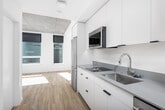 This single-floor space showcases a cohesive modern design defined by clean lines, a neutral white palette, and light wood-tone flooring that runs throughout. The layout connects functional areas in a streamlined fashion, with a well-appointed kitchen featuring white flat-front cabinetry extending to the ceiling, stainless steel appliances including a full-size refrigerator, an undermount sink, and integrated dishwasher. A stacked washer and dryer unit is conveniently positioned within the same corridor, maximizing efficiency.

The bathroom is designed with a minimalist aesthetic, incorporating a glass-enclosed shower, a wall-mounted white vanity with a gray stone countertop, an undermount sink, and a backlit mirror. Light wood-tone flooring continues here, adding warmth to the space.

The main living area benefits from multiple windows dressed with navy roller shades, allowing natural light to fill the room while maintaining privacy. An exposed concrete ceiling panel adds an industrial design element, contrasting with the otherwise bright and clean interior. A secondary room features a large window framing views of surrounding greenery and city skyline, further enhancing the connection to the outdoors. The entry corridor is accessed via a modern door with a smart lock, completing this thoughtfully designed urban space.