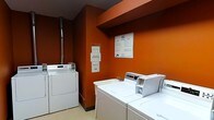 This shared laundry facility is defined by its striking burnt-orange walls, which give the utilitarian space a distinctive and energetic character. The room is equipped with multiple coin-operated washing machines and dryers arranged along the walls, providing ample capacity for resident use. The machines are positioned for easy access, with washers and dryers paired conveniently side by side. A vertical exhaust pipe runs along one wall, serving the dryer ventilation system. Informational signage and posted notices are displayed on the walls, offering guidance on machine operation, usage policies, and laundry hours. The flooring is a light-toned, easy-to-clean surface practical for a high-traffic utility space. The ceiling features a dark finish that contrasts with the vibrant wall color, adding depth to the room's overall aesthetic. The layout is straightforward and functional, prioritizing efficiency and ease of use. Overall, the space is a well-equipped, communal laundry room that balances practicality with a bold visual identity through its use of color.