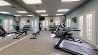 Located at 1301 Williamsburg Dr in Bossier City, Louisiana, this fitness center offers a comprehensive workout environment designed to support a variety of exercise routines. The single-floor space is thoughtfully laid out, featuring a full complement of cardio equipment including treadmills, stationary bikes, and elliptical machines from BH Fitness, positioned to maximize floor space and ease of movement between stations. A dedicated free weight area includes a multi-tiered dumbbell rack and a flat workout bench, catering to strength training needs. Large wall-mounted mirrors run along one side of the room, enhancing the sense of space and allowing for form checks during exercise. The room is finished with durable, patterned carpet tile flooring suited to high-traffic use. Soft teal accent walls add a fresh, energizing atmosphere, complemented by motivational word-art decals emphasizing focus, dedication, and commitment. Bright, recessed panel lighting ensures the space is well-lit throughout. Multiple glass-paned doors and windows along the walls invite natural light and provide views of the exterior, contributing to an open, airy feel. A wall-mounted television and a large industrial fan round out the practical amenities, making this fitness center a functional and inviting space for daily exercise.