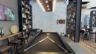 This single-floor entertainment space combines industrial aesthetics with a sophisticated lounge atmosphere. At the heart of the room sits a full-size billiards table with a sleek black felt surface, positioned beneath a striking overhead lighting fixture that illuminates the playing area with focused, ambient light. The whitewashed exposed brick walls lend a raw, urban character to the space, while the dark flooring and charcoal-toned accents create a refined contrast throughout.

Floor-to-ceiling built-in shelving units line both sides of the room, displaying an organized collection of books, decorative objects, and curated accessories that add warmth and visual interest. A dartboard mounted on the brick wall provides an additional recreational element, reinforcing the space's focus on interactive entertainment.

A small seating area with a round table and bar-height stools is tucked to one side, offering a comfortable spot for conversation or casual dining. The high ceiling enhances the sense of openness and volume, while the overhead pendant lighting contributes to the room's moody yet welcoming ambiance. Overall, this space is thoughtfully designed to serve as a versatile, upscale game room and social hub.
