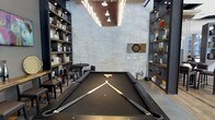 This single-floor entertainment space combines industrial aesthetics with a sophisticated lounge atmosphere. At the heart of the room sits a full-size billiards table with a sleek black felt surface, positioned beneath a striking overhead lighting fixture that illuminates the playing area with focused, ambient light. The whitewashed exposed brick walls lend a raw, urban character to the space, while the dark flooring and charcoal-toned accents create a refined contrast throughout.

Floor-to-ceiling built-in shelving units line both sides of the room, displaying an organized collection of books, decorative objects, and curated accessories that add warmth and visual interest. A dartboard mounted on the brick wall provides an additional recreational element, reinforcing the space's focus on interactive entertainment.

A small seating area with a round table and bar-height stools is tucked to one side, offering a comfortable spot for conversation or casual dining. The high ceiling enhances the sense of openness and volume, while the overhead pendant lighting contributes to the room's moody yet welcoming ambiance. Overall, this space is thoughtfully designed to serve as a versatile, upscale game room and social hub.