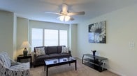 This single-floor space centers around a well-appointed living room designed for comfort and functionality. The room features soft, neutral-toned carpet flooring that adds warmth underfoot, complemented by clean, light-colored walls that create an open and airy atmosphere. A five-blade ceiling fan with integrated lighting serves as both a functional and decorative focal point overhead, ensuring year-round comfort and adequate illumination throughout the space. Large windows dressed with horizontal blinds line one wall, allowing generous amounts of natural light to filter into the room while maintaining privacy. The living area is thoughtfully arranged with a dark upholstered sofa accented by patterned throw pillows, a sleek black glass-topped coffee table, and a matching black media console, offering a cohesive and modern aesthetic. A bold black-and-white floral canvas print adorns the wall, adding an artistic touch that contrasts elegantly with the neutral surroundings. The overall ambiance is calm and inviting, with a balanced combination of natural light, neutral tones, and contemporary furnishings that make the space both practical and visually appealing.
