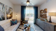 This single-floor residence presents a well-designed layout with a consistent contemporary aesthetic carried throughout each space. Light wood-look flooring runs continuously across the unit, complementing the crisp white walls and contributing to a clean, airy atmosphere.

The living area serves as the centerpiece of the home, featuring a ceiling fan with integrated lighting and a large sliding glass door framed by deep teal draping curtains. The door opens to an outdoor patio, allowing natural light to fill the room and providing views of the surrounding greenery.

The kitchen is thoughtfully appointed with white cabinetry, granite-style countertops, a subway tile backsplash, and black appliances. A peninsula with bar seating adds functionality and casual dining options, while pendant lighting and recessed fixtures illuminate the space effectively.

Both bedrooms are comfortably scaled, each benefiting from natural light through large windows fitted with blinds and sheer curtains. One bedroom includes an open closet with built-in shelving, offering practical storage solutions.

The bathroom features a long vanity with granite countertops, white cabinetry, a wide mirror, and a glass-enclosed walk-in shower, maintaining the clean and modern character found throughout the residence.