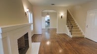 This two-story home offers a well-connected layout anchored by rich hardwood flooring that flows seamlessly throughout the main level. The living room serves as a welcoming focal point, highlighted by a classic white-mantled brick fireplace and a shuttered window that allows for controlled natural light. Wall sconces add a warm, ambient glow to the neutral-toned walls.

A graceful arched doorway draws the eye from the living area toward the dining space and into the kitchen beyond, where stainless steel appliances and white cabinetry are visible. A pendant light fixture hangs centrally within the archway, reinforcing the home's traditional character. The open sightlines between these spaces create a naturally flowing, functional layout ideal for everyday living and entertaining.

To the right, a staircase with elegant wrought iron railings and carpeted treads leads to the upper floor, adding a refined architectural detail to the entryway. The overall ambiance is warm and classic, combining traditional design elements — arched openings, decorative ironwork, and a brick fireplace — with clean, neutral finishes throughout.