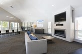 This single-floor community clubhouse, identified as Campus View, offers a well-appointed and thoughtfully designed gathering space for residents. The centerpiece of the facility is a generous open lounge area featuring soaring vaulted ceilings with clerestory windows that flood the interior with natural light. Comfortable seating arrangements, including plush sofas with accent pillows, a wall-mounted flat-screen television, and a sleek linear fireplace, create a welcoming and functional social environment. Durable carpeted flooring runs throughout the main lounge, complementing the neutral color palette of the walls and ceilings. A dedicated café-style kitchenette is outfitted with dark cabinetry, light stone countertops, stainless steel appliances, and a Starbucks coffee station, providing a convenient refreshment hub. Bar-height tables with modern stools offer casual seating in this area. Tall windows and a glass door in the kitchenette provide additional natural light and outdoor views. At the far end of the lounge, a bank of large windows and double French doors opens directly to an outdoor pool area, blurring the line between interior and exterior living. Accent seating near the pool-facing doors invites residents to relax and enjoy the surrounding views. The overall design strikes a balance between practicality and comfort.