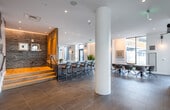 This single-floor amenity space is thoughtfully designed to blend modern aesthetics with functional versatility. The open-plan layout flows seamlessly between distinct zones, each offering its own character while maintaining a cohesive design language throughout.

At the heart of the space is a striking bar and kitchen area anchored by expansive marble countertops and dark navy cabinetry. Bar-height seating lines the counter, set against a dramatic dark brick accent wall illuminated by warm cove lighting. A slatted wood divider screen adds warmth and texture, partially framing the kitchen station equipped with a coffee bar and beverage area.

The lounge zone features generous white sectional sofas, a glass-top coffee table, and curated colorful artwork displayed against the rich dark brick wall. A large flat-screen television is mounted within a sleek dark built-in cabinetry unit, creating a comfortable media lounge environment.

A dedicated dining and conference area offers a long wooden table with plaid-upholstered seating beneath a linear pendant light fixture, complemented by bold photographic artwork on a deep charcoal accent wall. Elsewhere, a banquette seating nook with black leather upholstery and a slatted wood panel backdrop provides an intimate gathering spot.

Large glass entry doors and floor-to-ceiling windows draw in natural light, while recessed lighting, cylindrical white columns, and large-format tile flooring complete the polished, contemporary ambiance throughout.