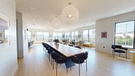 Located at 22 Mount Carmel Way in Worcester, MA, this two-level rooftop amenity space offers a sophisticated blend of indoor comfort and outdoor living. The generous main floor is bathed in natural light, thanks to a continuous wraparound arrangement of large, black-framed windows that frame sweeping views of the Worcester skyline. Light hardwood flooring runs throughout the interior, complementing the clean, contemporary aesthetic.

The indoor area is thoughtfully organized into distinct zones. A grand communal table anchored beneath striking oversized globe pendant lights serves as a central gathering point, ideal for large group meetings or social events. Adjacent lounging areas feature curated seating arrangements with a built-in fireplace set against a dramatic dark shiplap accent wall, creating an inviting and warm atmosphere. A dedicated bar area with a dark wood-paneled ceiling, pendant lighting, and counter-height seating adds a refined social hub to the space.

Ascending to the upper level, the open rooftop deck offers an expansive outdoor retreat finished in large-format pavers. It is outfitted with modular white lounge seating, sun loungers, a bar-height counter, and an outdoor television, all set against an unobstructed panoramic view of the city skyline and open sky above.