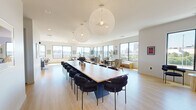 Located at 22 Mount Carmel Way in Worcester, MA, this two-level rooftop amenity space offers a sophisticated blend of indoor comfort and outdoor living. The generous main floor is bathed in natural light, thanks to a continuous wraparound arrangement of large, black-framed windows that frame sweeping views of the Worcester skyline. Light hardwood flooring runs throughout the interior, complementing the clean, contemporary aesthetic.

The indoor area is thoughtfully organized into distinct zones. A grand communal table anchored beneath striking oversized globe pendant lights serves as a central gathering point, ideal for large group meetings or social events. Adjacent lounging areas feature curated seating arrangements with a built-in fireplace set against a dramatic dark shiplap accent wall, creating an inviting and warm atmosphere. A dedicated bar area with a dark wood-paneled ceiling, pendant lighting, and counter-height seating adds a refined social hub to the space.

Ascending to the upper level, the open rooftop deck offers an expansive outdoor retreat finished in large-format pavers. It is outfitted with modular white lounge seating, sun loungers, a bar-height counter, and an outdoor television, all set against an unobstructed panoramic view of the city skyline and open sky above.
