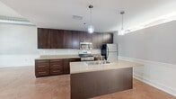 This single-floor space showcases a thoughtfully designed open layout that balances functionality with contemporary style. The kitchen serves as a central highlight, featuring rich dark-wood cabinetry extending to the ceiling, light stone countertops, and a full suite of stainless steel appliances including a gas range, built-in microwave, and refrigerator. A generously sized center island with an integrated sink is illuminated by two pendant lights, making it an ideal workspace and gathering point.

The living areas are defined by polished concrete floors in warm terracotta tones, lending an industrial yet refined character throughout. Large windows along one wall flood the main living space with natural light while framing views of surrounding urban architecture, creating a striking visual connection to the cityscape. Neutral gray walls accented by crisp white chair rail molding add a clean, layered dimension to each room.

The bathroom is accessed through a hallway flanked by dark-wood panel doors and features delicate mosaic tile flooring, a dark-wood vanity with a stone countertop, a soaking tub, and a well-placed mirror that reflects natural light. The overall ambiance of this space is modern and urban, with consistent design details that tie each area cohesively together.