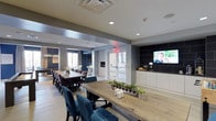 The Quincy Club Room is a thoughtfully designed communal space that seamlessly blends style and functionality. The open-plan layout accommodates multiple seating and dining configurations, anchored by two substantial live-edge and dark wood dining tables surrounded by plush velvet chairs in deep blue and eggplant tones, offering ample seating for large gatherings. Light hardwood flooring runs throughout, complementing the clean, contemporary aesthetic of the white and grey walls accented with bold dark panels. A wall-mounted flat-screen television is set against a striking dark tile backsplash, creating a dedicated entertainment zone alongside a sleek white cabinetry unit. A linear fireplace serves as a warm focal point, adding to the inviting ambiance of the space. Natural light filters in through glass-paneled entry doors and windows, brightening the interior and enhancing its welcoming atmosphere. Recessed lighting overhead ensures the space remains well-lit throughout the day. An adjacent area, partially visible and framed by an elegant chandelier and exposed brick wall, hints at additional lounge or dining functionality. The overall design strikes a balance between sophisticated and approachable, making it a versatile venue for both casual gatherings and formal events.