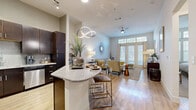 Located at 6215 Via La Cantera in San Antonio, TX, this single-story residence offers a thoughtfully designed layout that balances style and functionality. The open-concept living and kitchen area forms the heart of the home, where light wood-style flooring flows seamlessly throughout. The kitchen is appointed with rich dark-wood cabinetry, granite countertops, stainless steel appliances, a mosaic tile backsplash, and a central island with bar seating, illuminated by a striking modern pendant light and track lighting overhead. The living room extends naturally from the kitchen, bathed in natural light from large French doors that open to the outdoors. A ceiling fan and neutral palette create a relaxed, airy ambiance. The bedroom is a serene retreat, featuring plush carpet, large windows that invite natural light, and ample closet space supported by three dedicated closets. The bathroom is finished with tile flooring, granite-topped dark-wood vanity, a full soaking tub, and large-format wall tile. A dedicated laundry room with full-size washer and dryer hookups adds everyday convenience. Hallways connect the spaces efficiently, contributing to a well-organized and comfortable floor plan throughout.