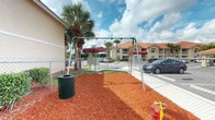 This outdoor playground area offers a welcoming recreational space designed for children within a residential community. The play area is surfaced with vibrant red wood mulch, providing a safe and cushioned ground cover beneath the equipment. A freestanding metal swing set serves as the centerpiece, accompanied by additional play elements including a small ride-on toy visible at the edge of the space. A dark green mesh waste receptacle is conveniently positioned nearby, reflecting attention to cleanliness and upkeep. The area is enclosed by a chain-link fence, clearly defining the play zone while maintaining open sightlines for supervision. A shaded canopy structure with a deep red awning sits toward the back of the space, offering a covered spot for rest. Tall, mature palm trees frame the area, lending a tropical character to the setting and providing natural shade. The surrounding residential buildings, featuring warm tan and yellow stucco exteriors with matching red-trimmed awnings and balconies, are visible in the background, tying the playground visually into the broader community. The space benefits from an abundance of natural light under a bright, open sky.