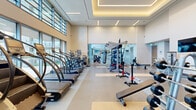 This well-appointed fitness center in Denver, Colorado offers a comprehensive exercise space designed to accommodate a wide range of workout routines. The main exercise area features soaring double-height ceilings with recessed tray lighting and large floor-to-ceiling windows that flood the space with natural light while providing views of the surrounding outdoors. The flooring transitions between durable tile and cushioned rubber matting, delineating zones for different training activities. A robust selection of cardio equipment lines the windowed perimeter, including treadmills and elliptical machines. The strength training area is well-stocked with Cybex cable machines, multi-functional weight racks, barbells, dumbbells, benches, and plate-loaded equipment, complemented by wall-mounted flat-screen televisions. A dedicated studio space is separated from the main floor by decorative etched glass partitions featuring a topographic map design, lending an artistic touch to the interior. This studio is finished with warm hardwood flooring, full-length mirrors, wall-mounted speakers, and a mounted television, and is equipped with spin bikes and step platforms. A hand sanitizer station and built-in storage cubbies enhance functionality throughout. Motivational wall graphics reinforce the athletic atmosphere, making this fitness center a dynamic and inspiring environment for all levels of exercise enthusiasts.