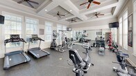 Located at 245 Creekwood Cove Ln in Lenoir City, Tennessee, this fitness center offers a thoughtfully designed exercise room that combines architectural elegance with practical functionality. The space is defined by its striking coffered ceiling, which adds visual depth and a polished aesthetic, while multiple ceiling fans ensure consistent air circulation throughout. Recessed lighting complements the abundant natural light streaming in through large, white-trimmed windows fitted with horizontal blinds, creating a bright and energizing workout environment. A full-length wall mirror visually expands the room and allows for form monitoring during exercise. The durable speckled rubber flooring provides cushioning and traction, making it well-suited for a wide range of physical activities. The room is comprehensively outfitted with cardio equipment including treadmills, a rowing machine, and a stationary bike, alongside strength-training options such as a multi-station cable machine, a dumbbell rack with an extensive free weight selection, medicine balls, and a weight bench. A wall-mounted television and a built-in water station add to the convenience of the space. The neutral, warm-gray wall tones and clean white trim throughout lend the room a contemporary and inviting atmosphere.