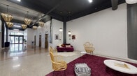 This single-floor space presents a thoughtfully designed lobby and lounge area that blends industrial architecture with vibrant, eclectic interior styling. The soaring dark-painted ceilings expose structural beams and columns, creating a loft-like atmosphere that feels both expansive and dynamic. Polished terrazzo flooring in the entry corridor — accented with warm terracotta inlays — transitions seamlessly into the broader lounge area, grounding the space with a classic yet contemporary feel.

A bold typographic mural reading "Let The Fun Begin" anchors one wall of the entry corridor, setting an inviting and energetic tone. A series of ornate gold pendant chandeliers and globe pendant lights hang at varying heights throughout, casting warm, ambient illumination across the space.

The lounge area is furnished with distinctive rattan fan-back chairs, a tufted olive-green velvet sofa, and a deep jewel-toned purple sofa, all arranged atop rich burgundy shag rugs. Gallery walls display a curated selection of framed artwork, adding a personalized and artistic character. A pair of classic elevator bays with brushed stainless steel and bronze-toned doors complete the functional elements of this welcoming space.