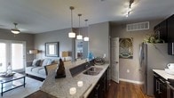 This thoughtfully designed single-floor residence showcases a seamless open-concept layout that connects the kitchen, dining, and living areas into one cohesive and functional living space. The kitchen serves as a central hub, featuring an expansive granite countertop island with a double stainless steel undermount sink, dark cabinetry, and a trio of sleek pendant lights that provide both task lighting and visual interest. A track lighting system overhead ensures the space remains well-lit throughout. The hardwood flooring runs continuously across the main living areas, adding warmth and a sense of continuity to the home's contemporary aesthetic. Neutral gray walls provide a sophisticated backdrop that complements the modern finishes found throughout. The adjoining living area is comfortably appointed, with a cozy seating arrangement anchored by a framed wall mirror that enhances the sense of depth and light. A doorway leads to additional areas of the home, hinting at further private spaces beyond. Decorative artwork adds personality and character to the interior. The overall ambiance is modern, clean, and refined, making this a welcoming and highly functional living environment.