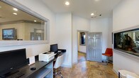 Located in Arlington, this single-floor space offers a clean, functional layout well-suited for professional use. The interior is finished with crisp white walls and warm stone-toned tile flooring that extends throughout, lending the space a polished yet approachable atmosphere.

The workstation area is equipped with a dark-surfaced desk, a desktop computer, and a printer, making it ideal for administrative tasks. A large pass-through window on one wall suggests a reception or service counter function, providing a clear line of communication between areas. Recessed lighting in the ceiling ensures consistent, even illumination across the workspace.

A gray paneled door near the workstation leads to an adjacent room, where a window allows natural light to filter in, enhancing the overall brightness of the space. An orange accent chair positioned nearby adds a subtle pop of color against the otherwise neutral palette. Security is supported by a visible ceiling-mounted camera, reflecting attention to safety within the environment.

The layout is efficient and well-organized, with clear sightlines and easy navigation between areas. Overall, the space presents a professional, welcoming environment designed to support day-to-day operational needs with clarity and comfort.