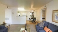 This single-floor residence is thoughtfully designed with an open-concept layout that seamlessly connects the living, dining, and kitchen areas into one cohesive and functional space. The living room features plush carpeting underfoot and is furnished with a comfortable seating area centered around a glass-topped coffee table, creating a relaxed and welcoming atmosphere. A ceiling fan overhead adds both style and practicality to the space.

The kitchen is positioned behind a half-wall breakfast bar, offering a sense of separation while maintaining visual openness throughout. It is equipped with white cabinetry, a white microwave, and a refrigerator, all contributing to a clean and bright aesthetic. The transition from the carpeted living area to hardwood flooring in the dining space adds a pleasant contrast in texture and warmth.

The dining area accommodates a compact table and chairs, well-suited for everyday meals. A window in the dining space allows natural light to filter in, enhancing the overall brightness of the interior. Adjacent to the main living area, a hallway leads to built-in shelving and closet storage, providing ample organizational space. The neutral wall tones and consistent design throughout give this residence a cohesive and inviting character.
