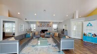 Located at 1700 Woodbury Rd in Orlando, FL, this single-floor community clubhouse offers a well-appointed gathering space designed with modern aesthetics and functional comfort in mind. The main lounge area is anchored by a striking floor-to-ceiling tiled fireplace, which serves as the room's focal point and is framed by decorative wood shelving and curated artwork. Soaring vaulted ceilings with recessed lighting contribute to an open, airy atmosphere, while warm hardwood-style flooring runs throughout, adding a sense of cohesion and warmth to the space. Contemporary gray upholstered sofas and armchairs are arranged around a glass coffee table atop a patterned area rug, accented with teal and gold throw pillows that echo the space's modern color palette. A cozy bistro-style seating nook with high-top chairs and a round glass table provides a more intimate setting near large windows that invite natural light. Adjacent to this nook, a dedicated coffee station offers a convenient self-serve setup with a single-serve brewer and condiment organizers. A separate workspace area with a desk and French doors leading outdoors rounds out the layout, making this clubhouse a versatile and inviting destination for residents.