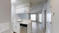 This single-floor space offers a clean, modern aesthetic characterized by an open layout that seamlessly connects the kitchen and living area. The kitchen is well-appointed with crisp white cabinetry — including upper and lower cabinets with sleek hardware — a stainless steel dishwasher, a double-basin sink with a contemporary faucet, and light-toned countertops that enhance the bright, airy feel. Overhead cabinetry extends above the kitchen peninsula, maximizing storage while maintaining an open sightline into the living area. The flooring throughout features light wood-look planks, adding warmth and continuity across the space. A textured ceiling adds subtle architectural character overhead. The living area is bathed in natural light, courtesy of large windows that frame views to what appears to be an outdoor balcony or patio, creating a welcoming and open ambiance. Neutral gray walls provide a versatile backdrop, complementing the light finishes throughout. The overall design is functional and modern, with a cohesive color palette of whites, grays, and natural wood tones that gives the space a fresh, move-in-ready appeal.