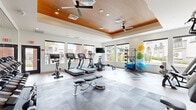 Located at 9600 W Georgia Ave in Glendale, Arizona, this fitness center is a thoughtfully designed, single-floor facility offering a comprehensive workout experience. The space is divided into two functional zones that together accommodate a wide range of exercise disciplines.

The architectural design is immediately striking, featuring a tray ceiling finished with warm wood paneling, recessed lighting, and dual ceiling fans that maintain comfortable airflow throughout. Large windows line the perimeter walls, flooding the interior with abundant natural light and offering views of the surrounding community. The flooring transitions between light hardwood-style planking and interlocking foam exercise mats, providing appropriate surfaces for both cardio and strength training areas.

The cardio zone is well-stocked with treadmills, elliptical machines, stationary bikes, and a rowing machine, all arranged to take advantage of the natural light and exterior views. A wall-mounted television is conveniently positioned for entertainment during workouts. The strength training area features a full multi-station cable machine, a dumbbell and kettlebell rack, a weight bench, and an extensive collection of stability and medicine balls displayed on a dedicated rack. Full-length mirrors along one wall visually expand the space and support proper form. The overall ambiance is clean, bright, and modern, making this fitness center a well-rounded amenity.
